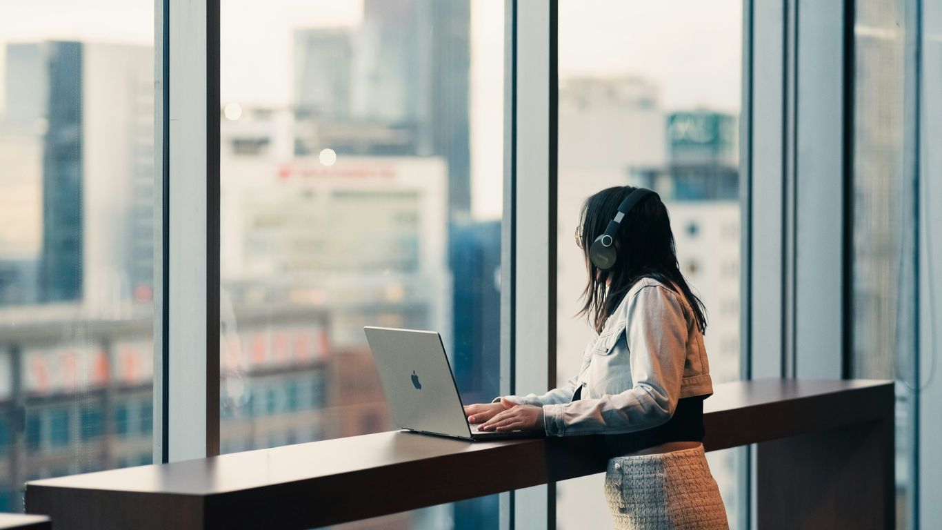 Woman wearing headphones works on laptop by window.